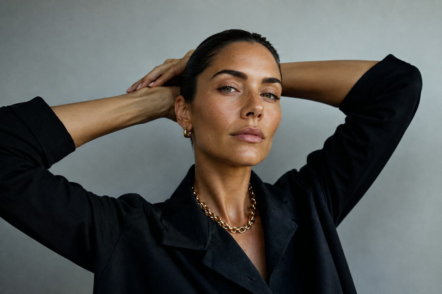 A woman in a black blazer with a gold chain, arms raised behind her head